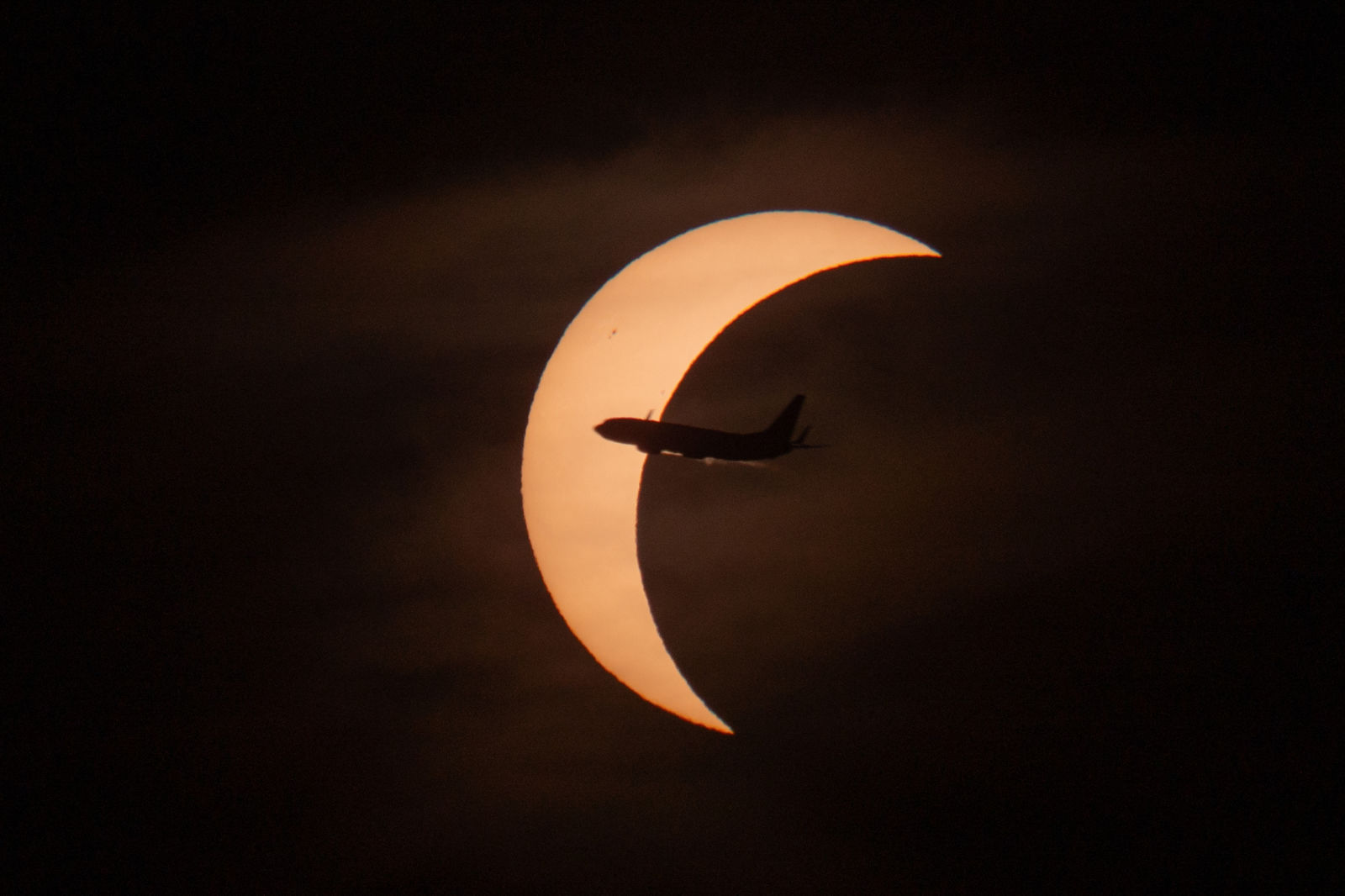 A crescent sun behind thin cloud during the partial phase, with an airliner silhouetted crossing it.
