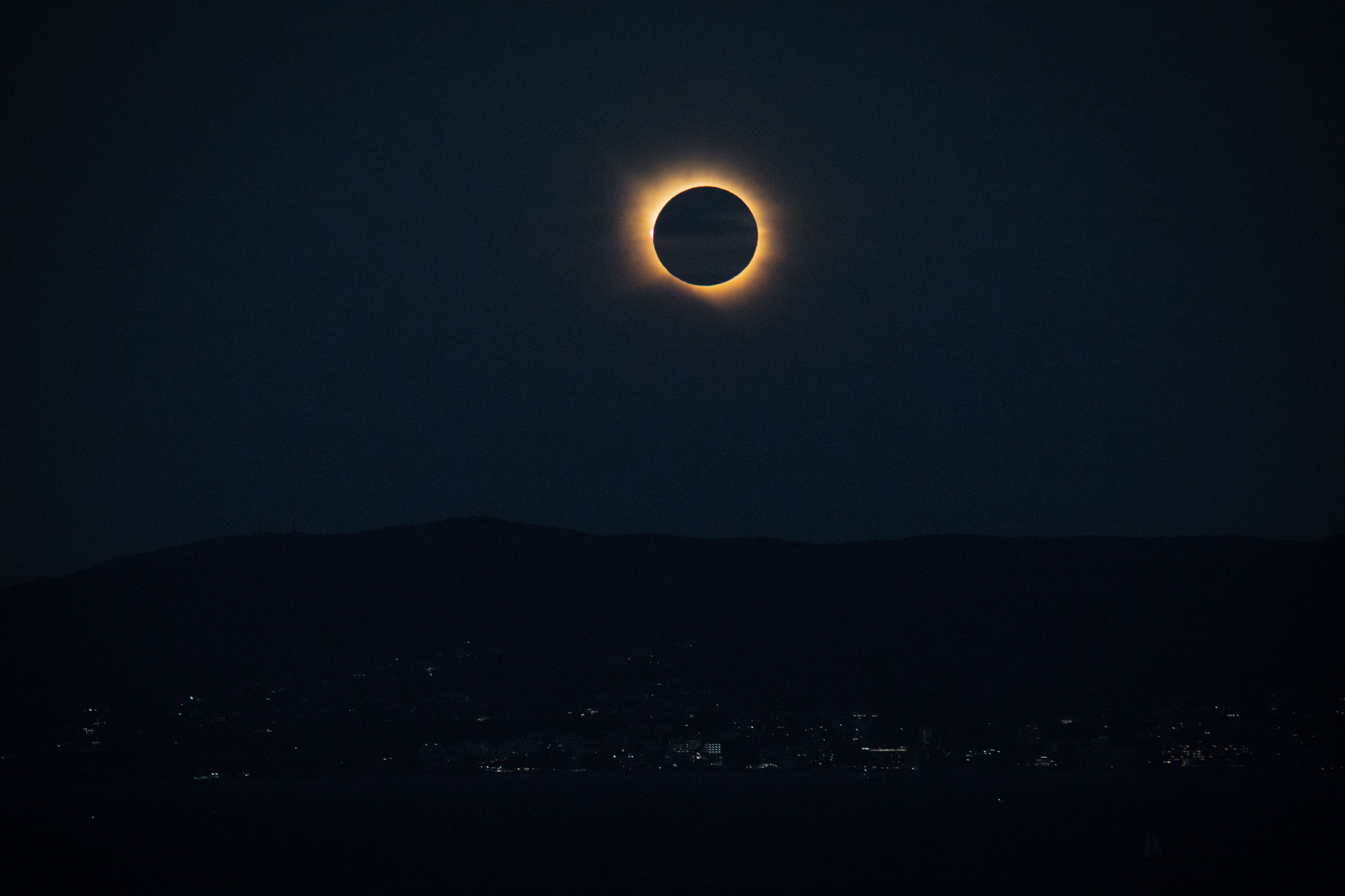 Wide view of totality low over the mountains across the bay, with the lights of the town coming on beneath it.