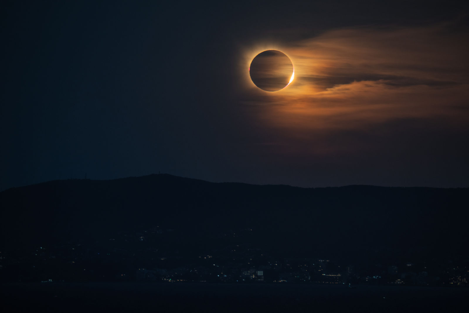 The diamond ring returning on the far side of the moon at the end of totality, low in orange-lit cloud above the mountains.