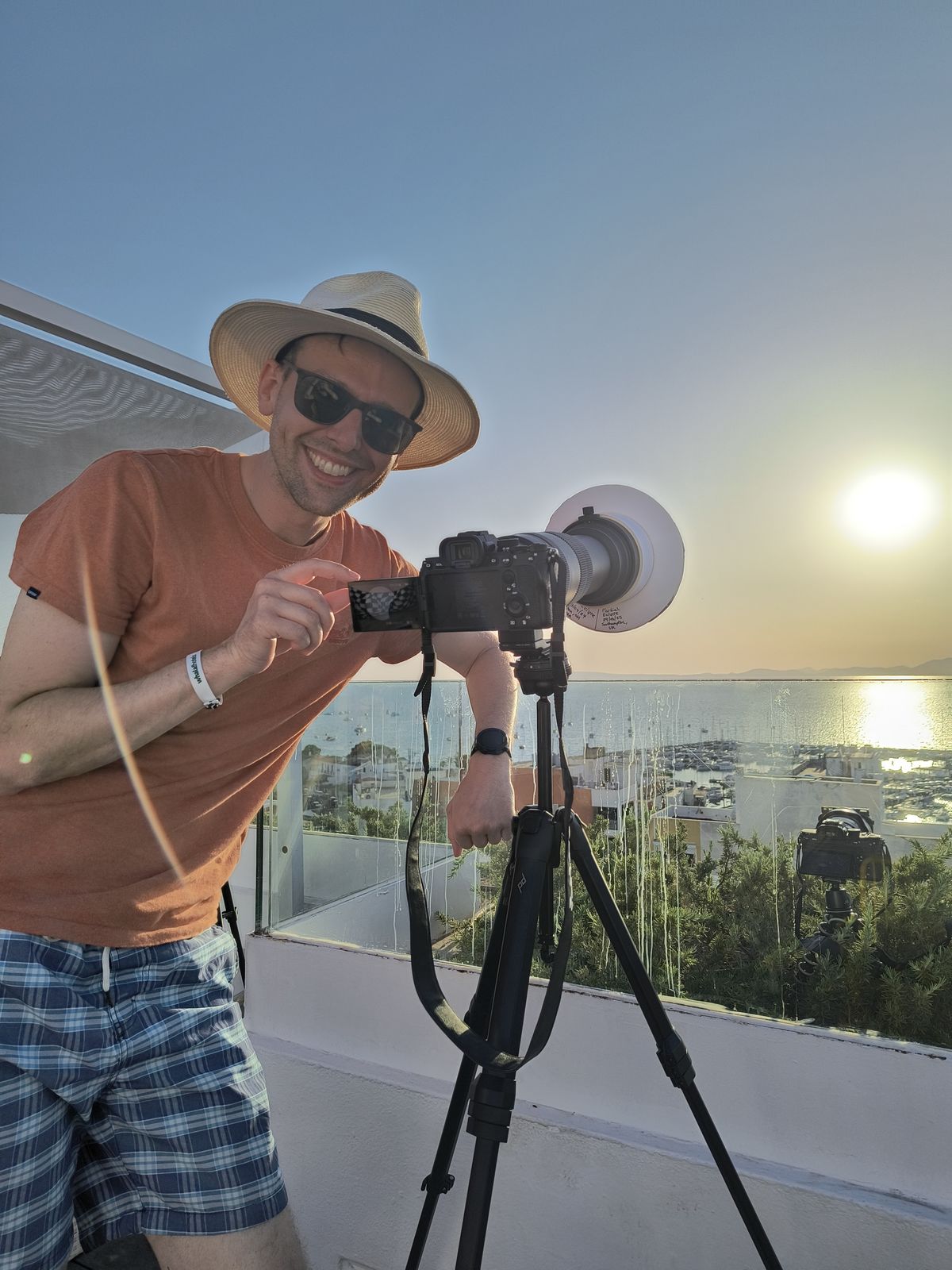Harry on the hotel roof beside a camera on a tripod fitted with a white solar filter, the bay behind him.