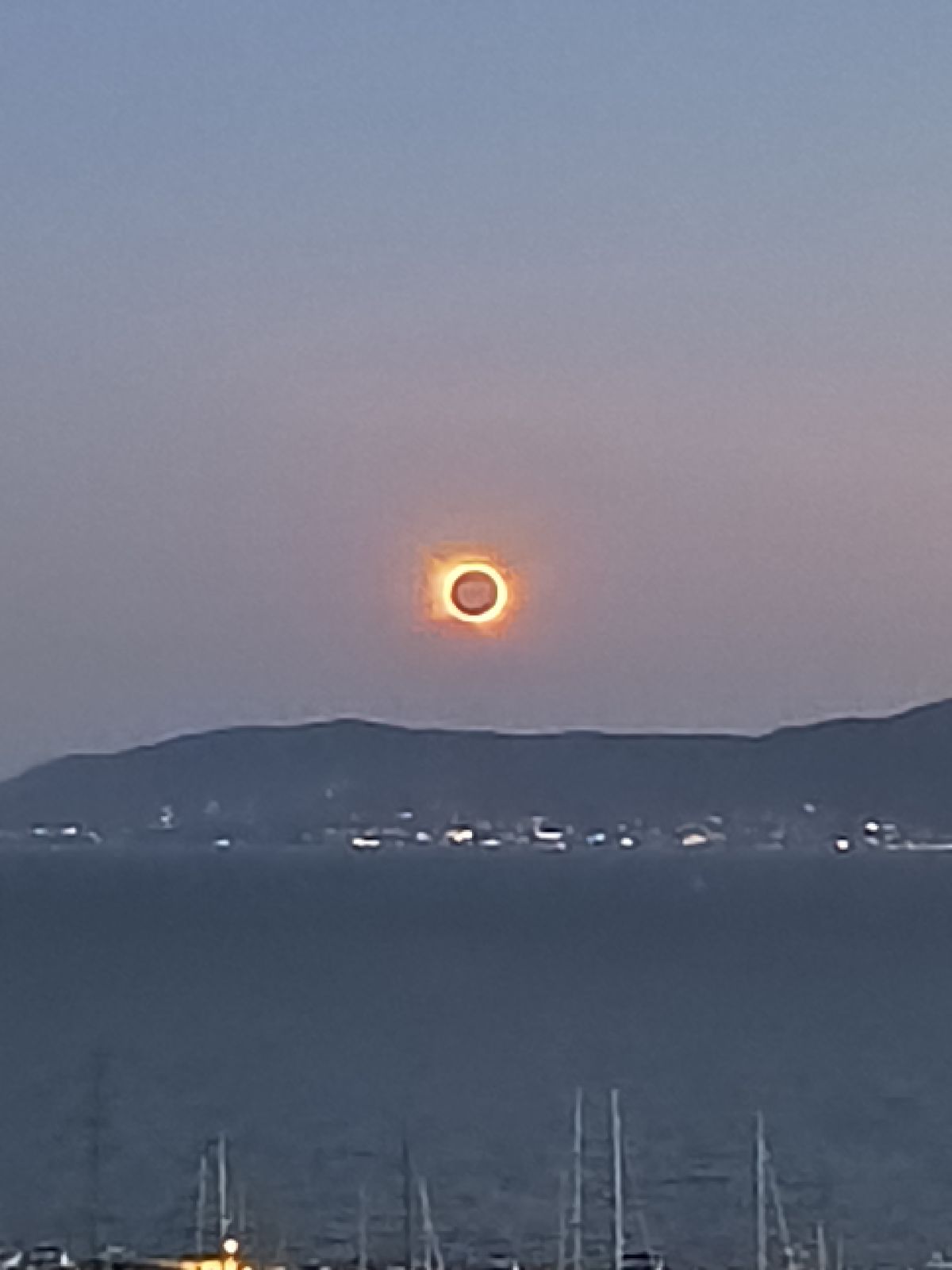 Totality seen from the hotel roof: a small bright ring above the mountains, with the lit marina below.